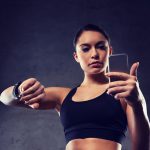 Mulher com top preto olha para o celular na mão esquerda e mantém mão direita erguida com relógio inteligente no pulso