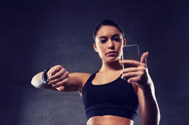 Mulher com top preto olha para o celular na mão esquerda e mantém mão direita erguida com relógio inteligente no pulso