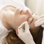 Mulher branca deitada na maca enquanto outra mulher de luvas faz suas sombrancelhas