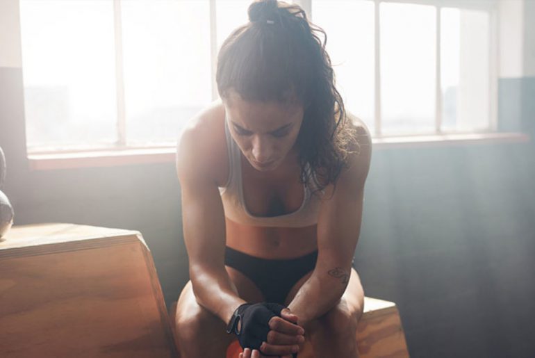 Mulher com cabelos presos cabisbaixa e ao lado e uma caixa de madeira e halter na academia