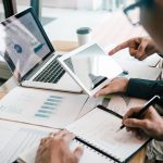 2 pessoas analisando algo no computador. Uma delas segura uma caderno e faz anotações