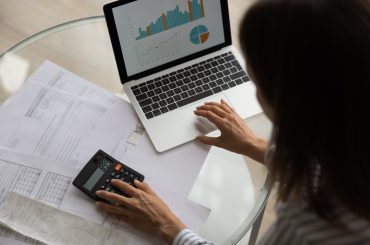 Mulher de cabelos pretos fotografada por cima digitando num notebook e usando calculadora