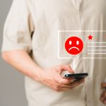 Imagem focalizada na mão de um homem mexendo no celular. Acima, uma ilustração de um emoji vermelho