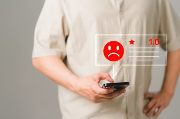 Imagem focalizada na mão de um homem mexendo no celular. Acima, uma ilustração de um emoji vermelho